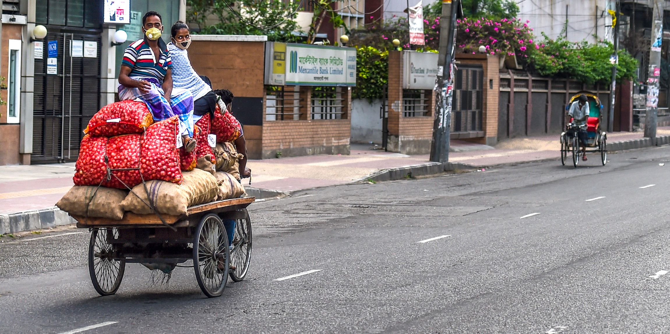 A man and a woman wearing facemasks sit on sacks of vegetables pulled by a three-wheeler rickshaw driver in Dhaka on April 18, 2020. | MUNIR UZ ZAMAN/AFP via Getty Images).