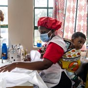 A woman in Nairobi sews protective clothing while carrying a child on her back.