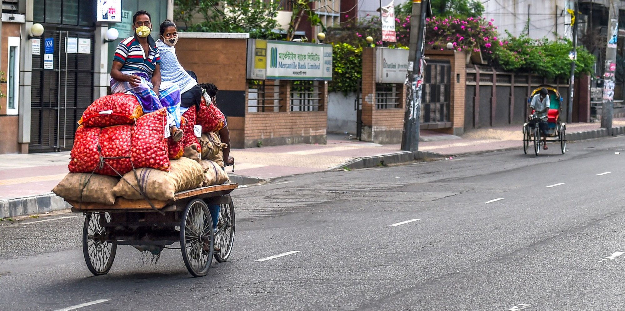 A man and a woman wearing facemasks sit on sacks of vegetables pulled by a three-wheeler rickshaw driver in Dhaka on April 18, 2020. | MUNIR UZ ZAMAN/AFP via Getty Images). 